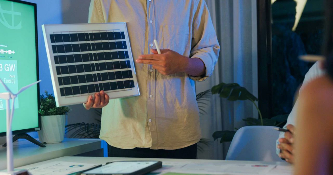 Un relatore tiene un piccolo modello di pannello solare durante una riunione, spiegando la tecnologia fotovoltaica e le strategie di gestione dei carichi a un gruppo.