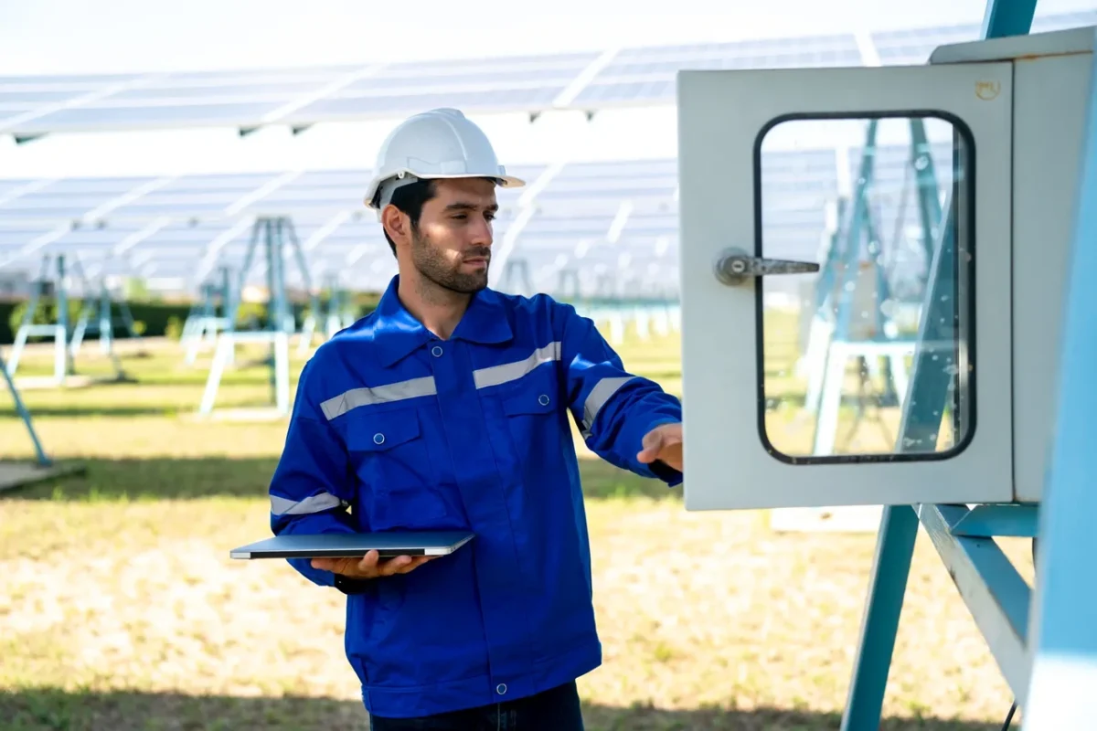 Ingegnere in blu apre l'armadio di controllo, monitorando i parametri dell'inverter per identificare guasti nel parco fotovoltaico.