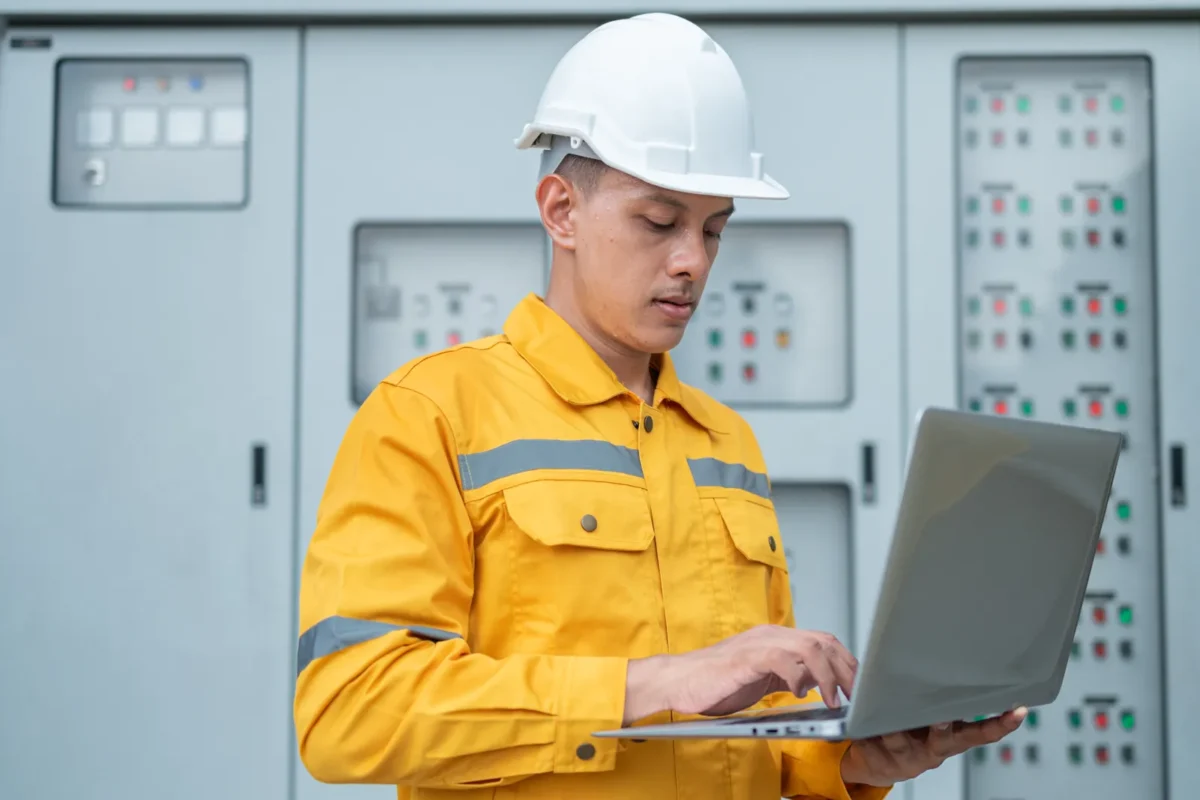 A power engineer in yellow workwear and white safety helmet operates a laptop to monitor and adjust hybrid inverter charger parameters at the electrical control cabinet.