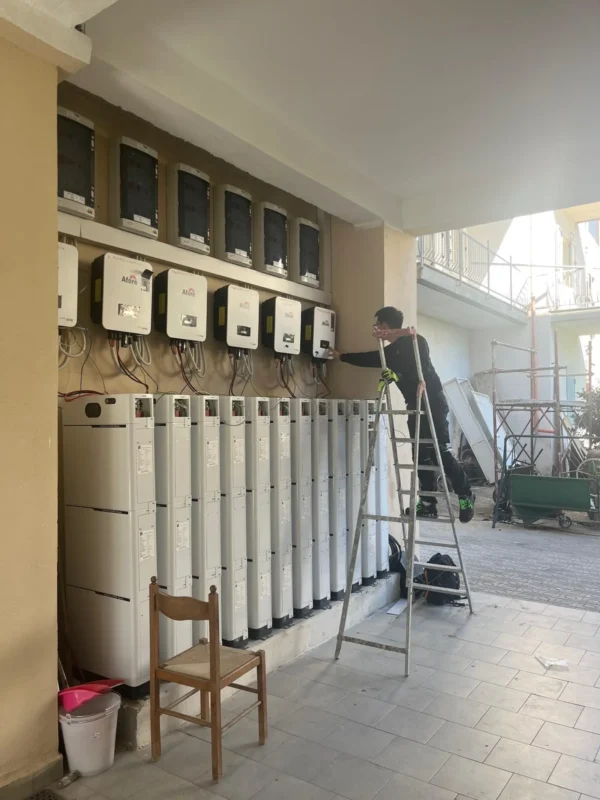 A technician stands on a ladder to inspect a row of mounted hybrid inverter chargers paired with stacked energy storage battery units outdoors.