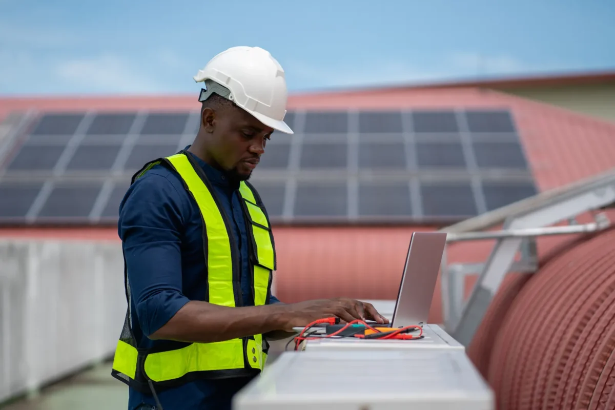 Solar technician with safety helmet and high-visibility vest uses a laptop to test rooftop solar panels paired with 48V 200Ah LiFePO4 battery storage units.