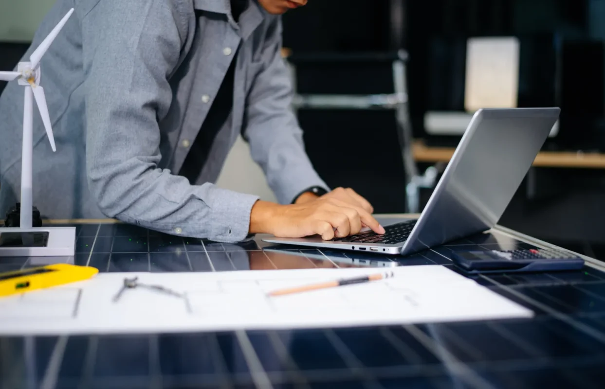 Renewable energy engineer works on a laptop to design solar storage schemes with wind turbine model and circuit drafts on solar panel patterned desk.