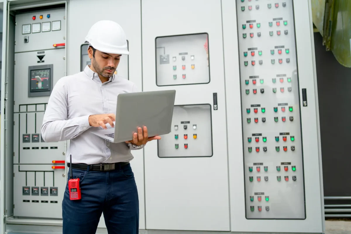 Electrical engineer wearing white safety helmet operates a laptop to monitor performance data of battery storage systems for commercial buildings at control panels.