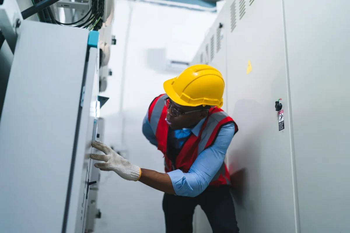 Electrical inspector wearing yellow safety helmet and reflective vest checks control cabinets of industrial commercial energy storage equipment indoors.