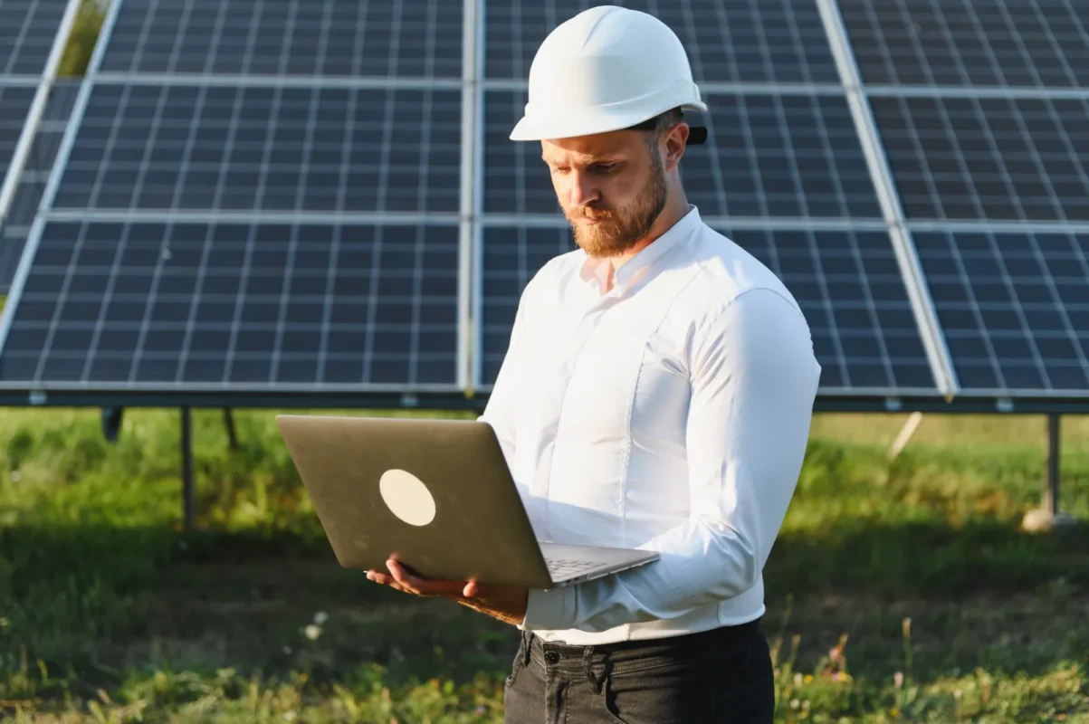 Solar engineer with white safety helmet uses a laptop to simulate power output and optimize commercial solar system design parameters on-site.