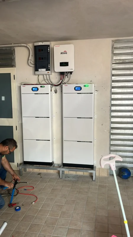 Electrician lays wiring on tiled floor to connect wall-mounted Afore inverter and two large vertical deep cycle lithium solar battery units indoors.